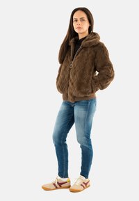 Giacca in pelliccia sintetica trapuntata marrone con cappuccio, chiusura con zip e tasche laterali, abbinata a jeans in denim blu e sneaker beige con accenti bordeaux.