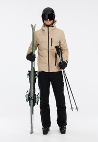 La tenue de ski comprend une veste rembourrée beige, un pantalon noir, des bottes de ski noires et des gants. Tient des skis verts et des bâtons de ski, portant un casque noir et des lunettes de ski.