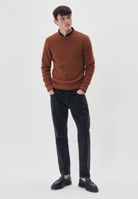 Suéter color óxido con puños y cuello acanalados, combinado con jeans negros y mocasines negros, de pie contra un fondo claro.