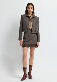 Blazer in tweed marrone con bottoni dorati, abbinato a una blusa a fantasia e a una gonna a tubo in tweed coordinata con pizzo. Scarpe borgogna e calzini alla caviglia.