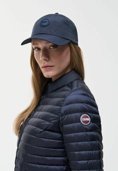 Woman wearing a dark blue quilted jacket with a Colmar logo on the sleeve and matching Colmar baseball cap, looking toward the camera.