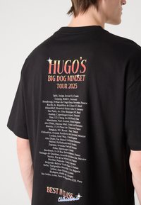 Černé bavlněné tričko s barevným nápisem "Hugo's Big Dog Mindset Tour 2025" na zadní straně, které uvádí více městských lokalit.