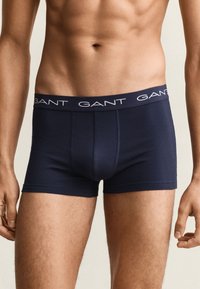 Torse masculin portant un boxer bleu marine avec le logo GANT sur la ceinture, debout, les bras détendus le long du corps, sur un fond uni.
