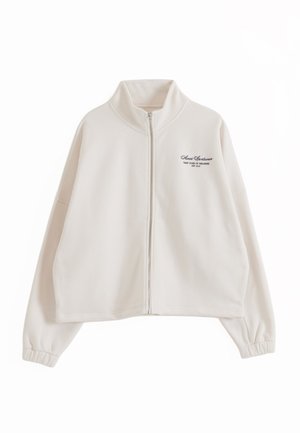 Giacca beige con zip, collo alto, polsini elastici e piccolo logo testuale nero sul petto sinistro con la scritta "Almond Sportswear."