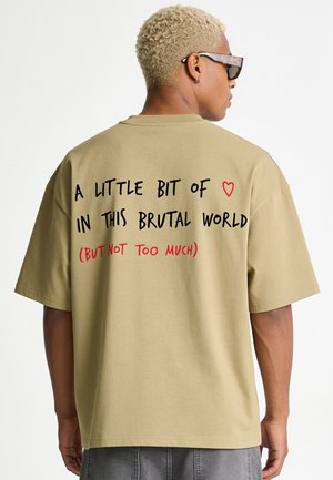 Personne aux cheveux blonds courts et lunettes de soleil portant une chemise beige avec le texte "A little bit of love in this brutal world (but not too much)" dans le dos.