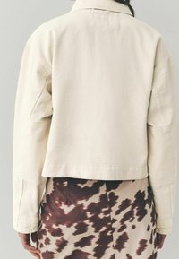 Beige cropped jakke med klassisk krave, lige snit og struktureret stof, kombineret med en brun og creme-mønstrede nederdel.
