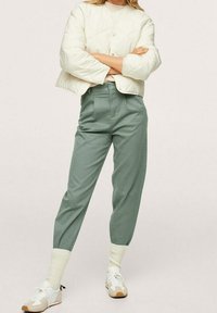 Kobieta w kremowej pikowanej kurtce, szałwiowych spodniach z wysokim stanem, kremowych skarpetach prążkowanych oraz beżowo-białych sneakersach, stojąca z założonymi na piersi rękami.