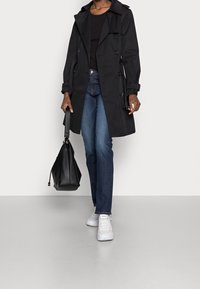 Černý trenčkot s páskem v pase, knoflíky a klasickým límcem, spárovaný s modrými džíny a bílými teniskami. Nese černou kabelku.