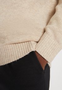 Beige ullgenser med ribbestrikket kant, med en sideslitt og teksturert strikk. Kombinert med svarte tøybukser.