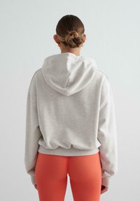 Mujer con el cabello recogido en un moño, que lleva pendientes de aro dorados, un sudadera gris claro y leggings de color naranja brillante, de pie contra un fondo blanco liso.