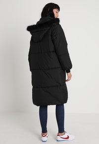 Manteau en duvet noir avec capuche, doté d'une bordure en fausse fourrure, de poignets élastiques et d'un design long et oversize. Porté avec un jean bleu et des baskets.