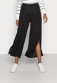 Pantalons larges noirs avec taille à cordon de serrage, présentant des fentes avant et un tissu léger et texturé. Associés à des baskets blanches.
