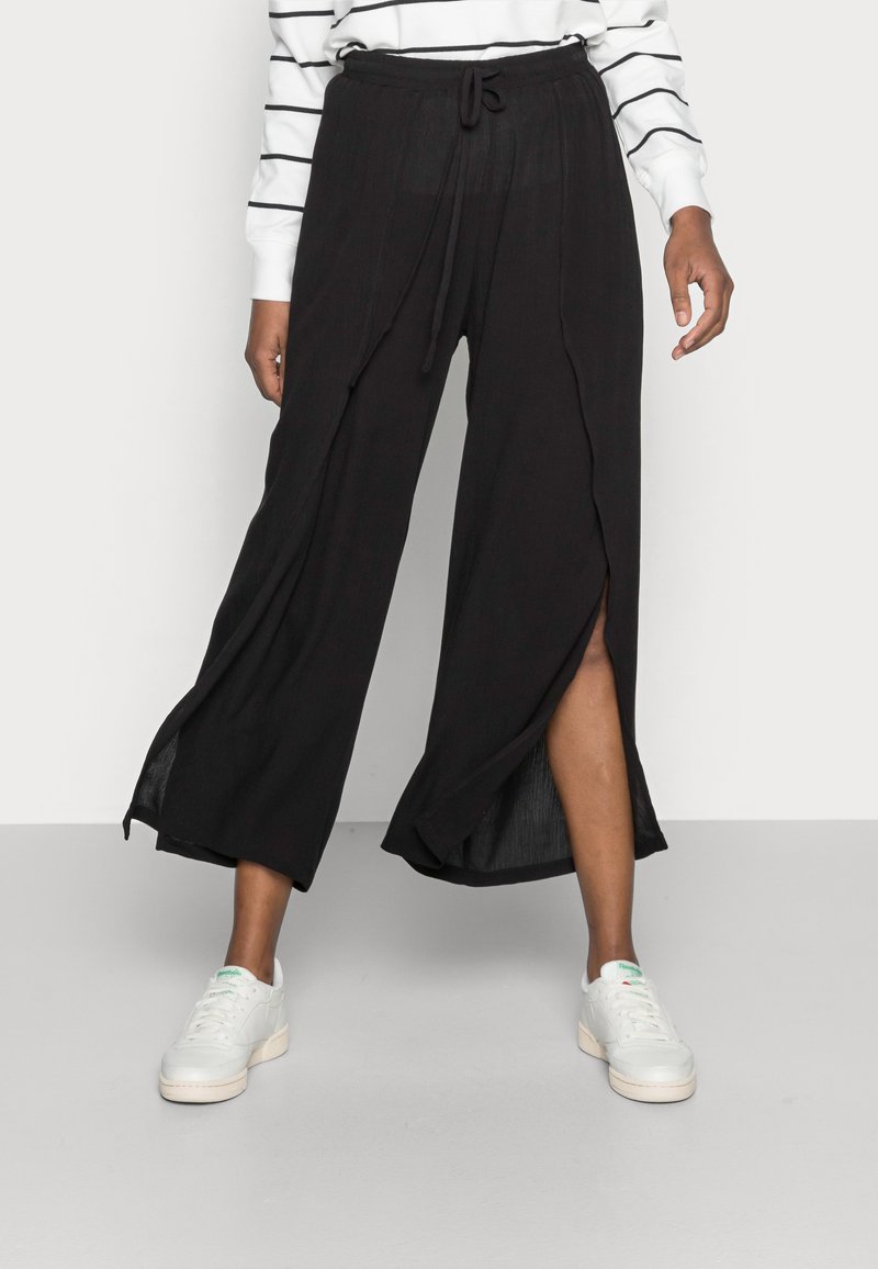 Pantalons larges noirs avec taille à cordon de serrage, présentant des fentes avant et un tissu léger et texturé. Associés à des baskets blanches.
