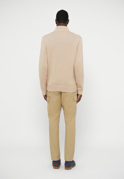 Beige kötött pulóver magas gallérral, bordás mandzsettákkal és derékpánttal, khaki cargo nadrággal és színes sportcipőkkel párosítva.