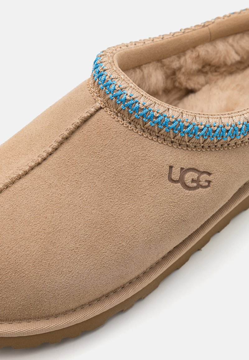 Tijdloze suède pantoffels in taupe met een gevoerd interieur, een blauw en beige geborduurd accent rondom de kraag en een rubberen loopzool. UGG-logo aanwezig.