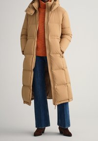 Lange tan pufferjas met een capuchon, voorzien van een ritssluiting aan de voorkant, een gequilte uitstraling en zijzakken. Draag het over een gestructureerde oranje trui en blauwe jeans.