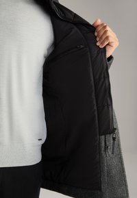 Grau melierter Wollmantel mit einer schwarzen Steppfutter. Verfügt über eine mit Reißverschluss verschlossene Innentasche und eine strukturierte Außenseite. Hell gefärbter Pullover darunter.