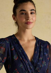 Tumesinise lillemustriga pluus punase, sinise ja rohelisega, millel on puhvsete varrukate ja V-kaelus. Valmistatud kergest, läbipaistvast kangast.