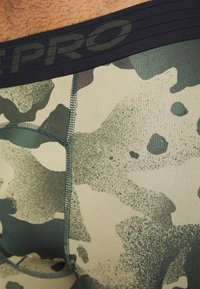 Gros plan sur un tissu camouflage vert et beige avec une ceinture élastique noire marquée "PRO" à la taille d'une personne.