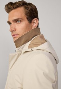 Chaqueta impermeable beige con un alto cuello, que presenta un acento marrón claro en el cuello. Textura suave con un diseño minimalista y cremallera frontal.