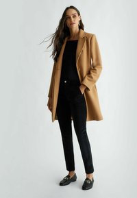 Cappotto in misto cammello, top aderente nero, pantaloni in corduroy neri, mocassini neri con dettagli in oro. Texture liscia e vestibilità sartoriale.