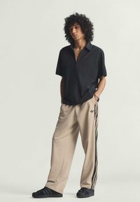 Čierna krátka zipsová košeľa s krátkym rukávom, béžové široké nohavice s čiernymi pruhmi a čierne tenisky. Voľný strih, hladká textúra, minimálny detail loga.