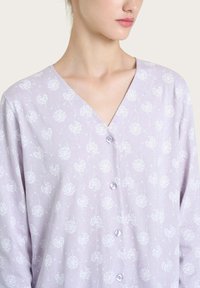Camicia lilac a bottoni con scollo a V, caratterizzata da un motivo di dente di leone bianco. Realizzata in un tessuto morbido con una trama leggera e liscia.