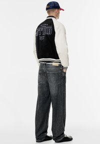 Veste varsity noire et crème avec "WELCOME TO STUD" brodé dans le dos, associée à un jeans en denim gris foncé à coupe ample et des chaussures noires.
