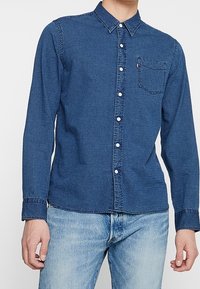 Muž v tmavě modré džínové košili s dlouhým rukávem a zapínáním na knoflíky, s kapsou na hrudi, stojící na jednobarevném pozadí, v světlých modrých džínách.