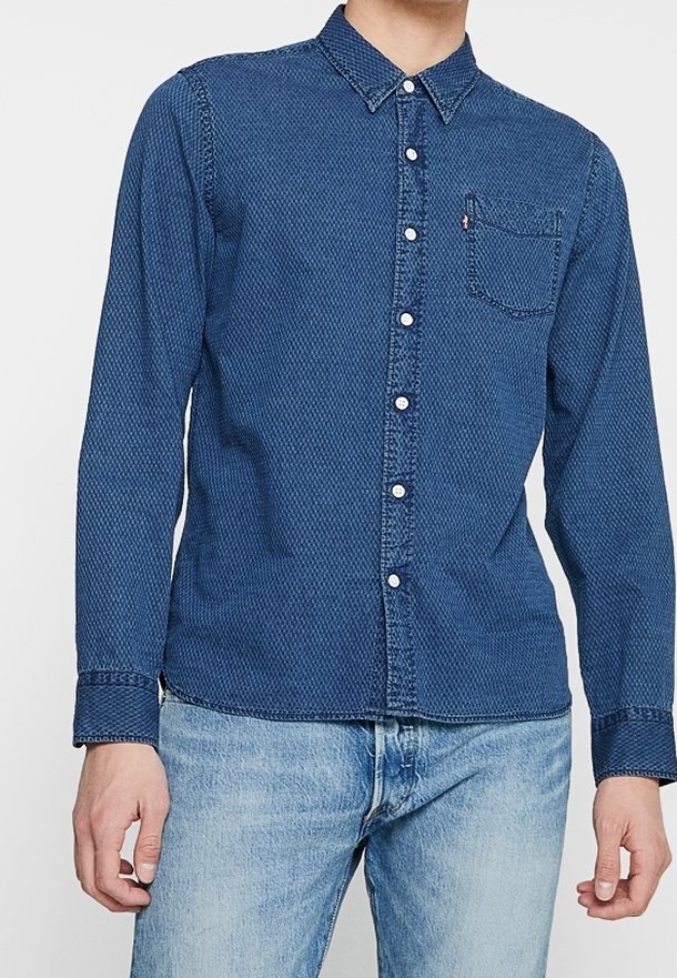 Mężczyzna w długim rękawie, zapinanej na guziki, ciemnoniebieskiej koszuli dżinsowej z kieszenią na piersi oraz jasnoniebieskich dżinsach, stojący na jednolitym tle.