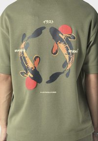 Khaki t-shirt z grafiką dwóch ryb w kolorach czarnym, pomarańczowym i czerwonym, z białym napisem "IRASUTO" oraz "ILLUSTRATED STUDIOS". Krótkie rękawy.