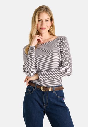 Blonde Frau mit langen Haaren trägt ein gestreiftes Langarmshirt, blaue Jeans mit braunem Gürtel, posiert mit der Hand am Gesicht vor weißem Hintergrund.
