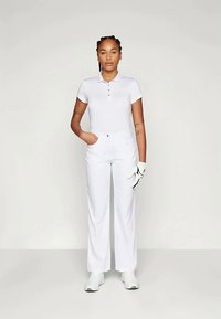 Sieviete stāv, ģērbusies pilnīgi baltā tērpā ar polo krekliņu, biksēm, baltām kurpēm un baltu cimdu labajā rokā, uz vienkārša gaiša fona.