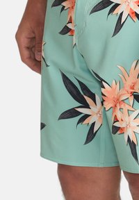 Kvetinové plavky v mätovo zelenej farbe, s broskyňovými a čiernymi vzormi kvetov, vyrobené z jemného a ľahkého materiálu s prešitým lemovaním.