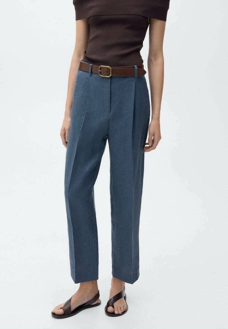 Femme portant un pantalon bleu taille haute avec une ceinture marron, un haut marron foncé épaules dénudées et des sandales noires à bout ouvert, debout devant un fond blanc.