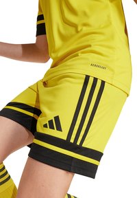 Shorts de sport jaunes avec des rayures noires, marque Aeroready et une coupe ajustée. Tissu texturé et accents noirs au niveau de l'ourlet.