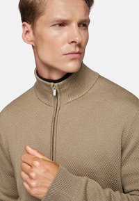 Beige zip-neck sweater with a textured knit, ribbed collar, and cuffs. Zipper pulls are silver-toned. Model's hand rests on chest.