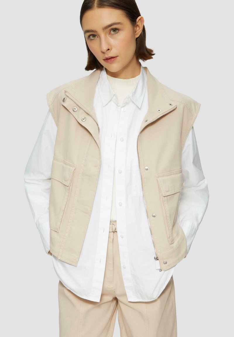 Gilet sans manches beige avec fermeture éclair et boutons pression, deux poches avant, superposé sur une chemise blanche à boutons, avec une texture lisse.