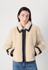 Crème-kleurige faux fur jas met zwarte leren afwerking, ritssluiting en verstelbare manchetten. Voorzien van zijzakken en een kraag.