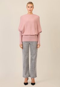 Hellrosaner, gestrickter Pullover mit geripptem Muster und hohem Kragen, kombiniert mit grauen, gemusterten, weit geschnittenen Hosen und schwarzen spitzen High Heels.