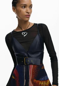 Top nero trasparente a maniche lunghe sotto un vestito di pelle blu navy con scollo a V, vita cintata e gonna con vivace motivo grafico. Collana d'argento.