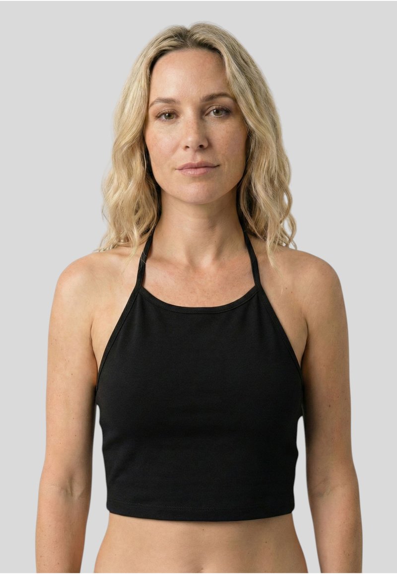 Femme aux cheveux blonds ondulés mi-longs, portant un crop top noir sans manches à col halter, debout devant un fond gris uni.