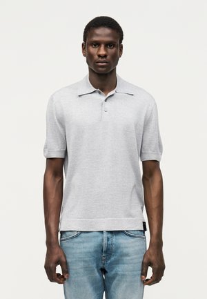 Άνδρας που φοράει ανοιχτογκρι πόλο με κοντά μανίκια και ανοιχτό μπλε τζιν, στέκεται μπροστά σε απλό λευκό φόντο.