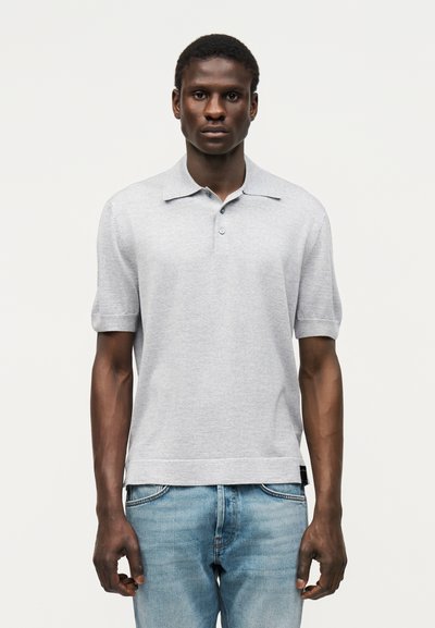 Homme portant un polo gris clair à manches courtes et un jean bleu clair, debout devant un fond blanc uni.