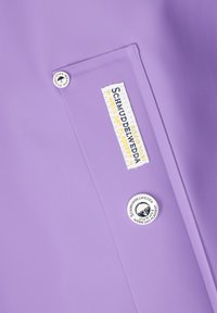 Violetinė vandeniui atspari medžiaga su kišene ir dviem apvaliais metaliniais akcentais. Yra siuvinėta etiketė su užrašu "SCHMUDDELWETTER" geltonos ir juodos spalvų.