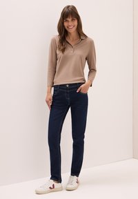 Mujer con cabello largo castaño sonríe, vistiendo un polo de manga larga beige, jeans azul oscuro y zapatillas blancas con detalles rojos.