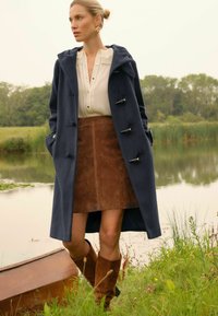 Cappotto in misto lana blu navy con cappuccio e chiusure a toggle, abbinato a una gonna in suede marrone e stivali alti in suede marrone. Sfondo naturale verde.
