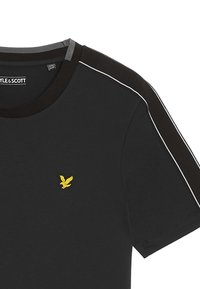 T-shirt nera con scollo rotondo, con accenti grigi e marroni sulle spalle e un piccolo logo di un uccellino giallo ricamato sul petto.