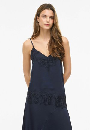 Mladá žena s dlhými hnedými vlasmi, ktorá má na sebe tmavomodré šaty bez rukávov s čipkovými detailmi na hrudi a leme, stojí na bielom pozadí.