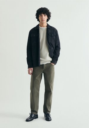 Młody mężczyzna z kręconymi włosami stoi, ubrany w czarną kurtkę, beżową koszulę, oliwkowe spodnie i czarne buty na jednolitym jasnym tle.